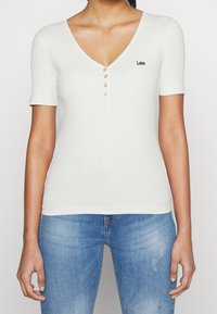Bílý žebrovaný top s krátkým rukávem, s hlubokým výstřihem do V, vpředu s pěti knoflíky a malým vyšitým logem "Lee" na levé části hrudi.