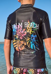 Juodas apsauginis marškinėlis su ryškiais, daugiaspalviais tropinių lapų raštais ir užrašu „PALM BEACH Florida“ gale. Lygi, blizgi medžiaga.