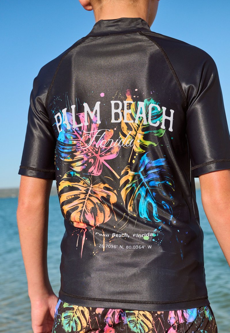 Schwarzes Rashguard mit lebhaften, mehrfarbigen tropischen Blattmotiven und dem Text "PALM BEACH Florida" auf der Rückseite. Glattes, glänzendes Material.
