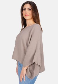 Hellbraunes, strukturiertes Poncho-Top mit weiten Ärmeln und Spitzenbesatz am Saum, kombiniert mit eng anliegenden blauen Jeans.