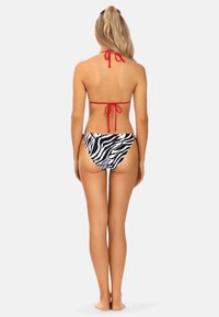 Czerwony biustonosz typu halter z zawiązywaniem z tyłu, zestawiony z białymi i czarnymi majtkami kąpielowymi w zebrę. Oferuje minimalne zasłonięcie i jest wykonany z elastycznego materiału.