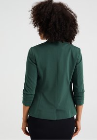 Tummanvihreä istuva bleiseri, jossa on kolme neljännesosaa hihat, sileä pinta ja takakappaleen tuuletus. Yksinkertainen muotoilu, ei näkyviä metalliosia tai kuvioita.