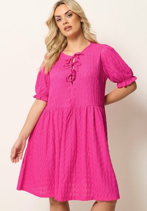 Blondynka ubrana w jasnoróżową teksturowaną sukienkę z krótkimi bufiastymi rękawami i trzema związanymi kokardami przy dekolcie, pozująca na jednolitym tle.