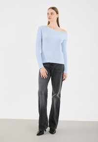 Donna che indossa un maglione a coste blu chiaro con spalle scoperte, jeans neri sbiaditi a gamba dritta e décolleté neri a punta, in piedi su uno sfondo neutro.