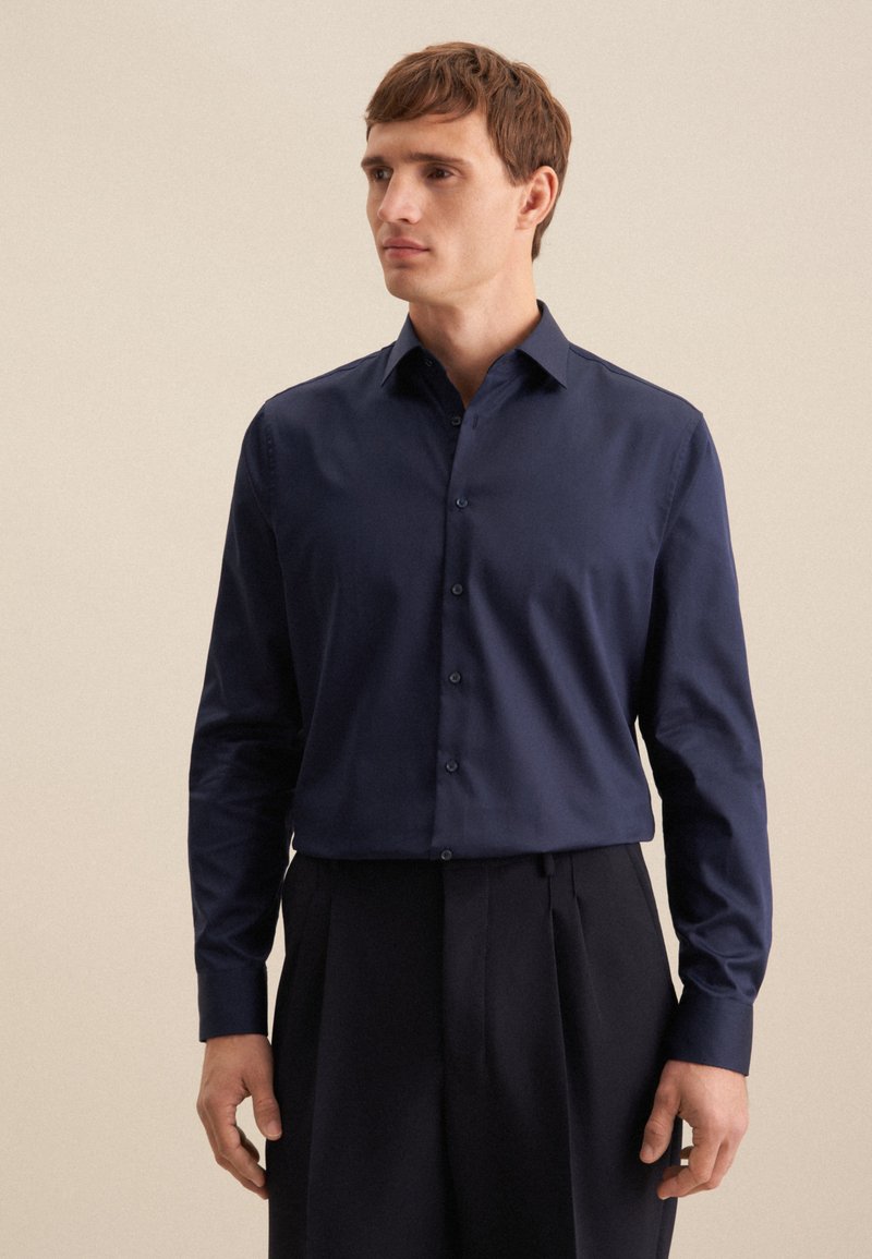 Chemise button-up bleu marine en tissu lisse, avec un col pointu et des manches longues, associée à un pantalon plissé sombre.