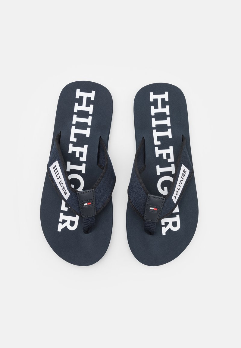 Mørkeblå flip-flops med hvid "HILFIGER" tekst på toppen, stofstropper og et logo mærke, med en flad sål og minimalistisk design.