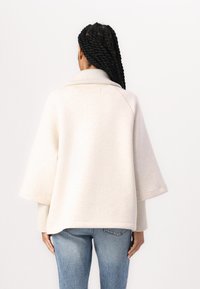 Lys beige oversized sweater med krave og brede ærmer, lavet af blødt stof, med ribbet tekstur og diskret logo på ryggen.