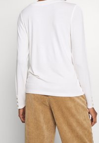 Blusa blanca de manga larga con escote redondeado y detalles de botones en los puños, combinada con pantalones de pana beige.