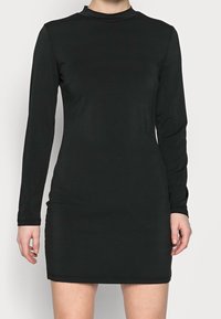 Mustavilla pitkähihaisella mekolla on sileä kangas, korkealle ulottuva kaulus ja tiukka siluetti, joka päättyy polven ylle.