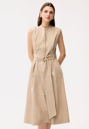 Vrouw met lang bruin haar, gekleed in een mouwloze beige midi-jurk met ceintuur en gouden knopen, en bijpassende gouden oorringen, staand.