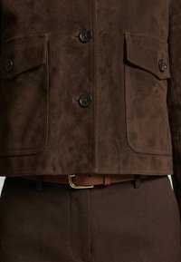 Brun ruskindjakke med knaplukning på klaplommer, båret over matchende brune bukser og et brunt læderbælte med guldlås.