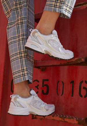 Personne portant des baskets New Balance blanches et argentées et un pantalon à carreaux bleu marchant sur des escaliers métalliques rouillés de couleur rouge.