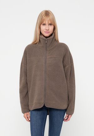Brun fleecejakke med høj krave, fuld lynlås og oversized pasform. Har en tekstureret overflade og raglanærmer, parret med blå jeans.