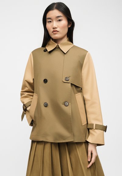 Jonge vrouw die een tweekleurige beige en zandkleurige dubbelgeknoopt trenchcoat draagt met gespen aan de manchetten en een geplooide rok.