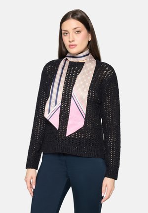 Frau trägt einen schwarzen Strickpullover, marineblaue Hose und ein rosa gemustertes Seidentuch, das um ihren Hals gebunden ist, vor einem schlichten Hintergrund.