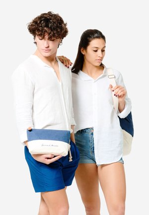 Blaue und cremefarbene Canvas-Tasche mit Reißverschluss und Quaste, auf der das Logo "Garrazzo Amarte" prangt. Modelle tragen legere Kleidung und halten die Tasche.