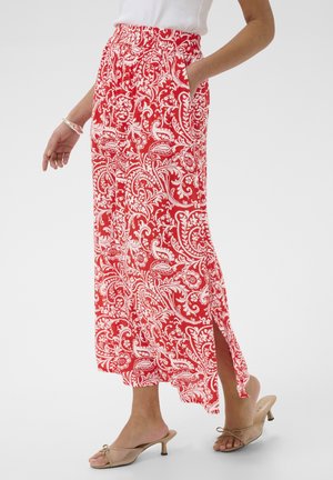 Žena v červeno-bielej maxi sukni s kvetinovým vzorom a rozparkom na boku, s jednou rukou v vrecku, v páre s béžovými sandálmi na nízkom opätku.