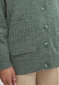 Zelený pletený svetr s žebrovanými kapsami, pěti velkými knoflíky a měkkou texturou. Tkanina vypadá teple a příjemně.