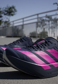 Chaussures de sport noires et roses posées sur du béton avec une rambarde métallique et des arbres flous en arrière-plan sous un ciel dégagé.