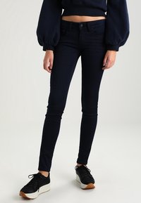 Mörkblå smala jeans i stretchmaterial, med en klassisk femfickdesign och metallknäppning, kombinerat med svarta sneakers.