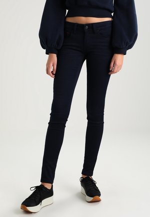 Jeans skinny azul marino en tejido elástico, con un diseño clásico de cinco bolsillos y cierre de botón metálico, combinados con zapatillas negras.