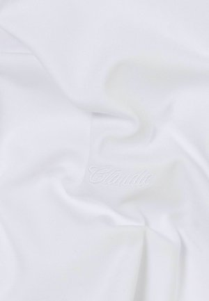 Donna che indossa una camicia bianca a maniche corte, pantaloni marroni a gamba larga, scarpe color granata, foulard blu fantasia in testa e tiene una borsa nera a tracolla.