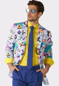 Blazer colorato con motivi geometrici in rosa, blu e giallo; camicia gialla; cravatta blu; pantaloni blu; occhiali da sole riflettenti.