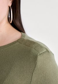 Oliven grønn strikket skjorte med rund hals og subtile detaljer på skuldrene. Stoffet ser mykt ut med en jevn tekstur.