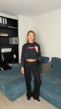 Czarna krótka bluza z różowym nadrukiem, w zestawie z czarnymi rozszerzanymi spodniami. Ciemne trampki. Stoi przed niebieską kanapą. Gładkie tło.