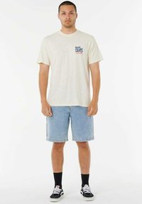 Krótki beżowy T-shirt z małym niebieskim i czerwonym logo "Rip Curl", w zestawie z jasnoniebieskimi dżinsowymi szortami i czarnymi skarpetami oraz białymi trampkami.