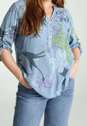 Kobieta nosząca jasnobeżową kwiecistą bluzkę z podwiniętymi rękawami i niebieskie dżinsy, ręce w kieszeniach, na jednolitym tle.