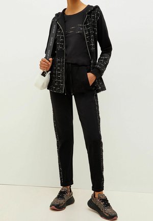 Sort hættejakke med teksturerede detaljer og lynlås, parret med sorte joggers med en sidestribe og sorte sneakers med et mønstret design.
