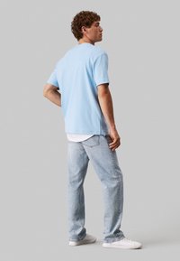 T-shirt di cotone azzurro chiaro con maniche corte, vestibilità comoda e scollo rotondo, indossata sopra una maglietta bianca, abbinata a jeans azzurri chiari e sneakers bianche.