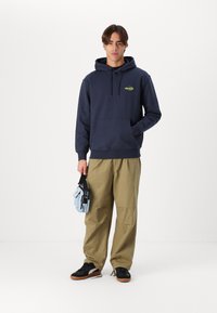 Navy hoodie with a front pocket and green logo, paired with loose khaki pants and black sneakers. Holds a light blue bag.