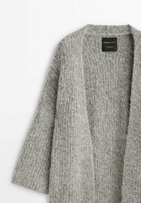 Cardigan doux et gris en maille duveteuse avec ouverture frontale, manches longues et une étiquette noire « Massimo Dutti Studio » à l'intérieur du col.