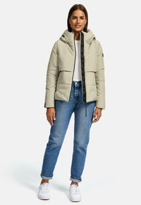 Lichtbeige pufferjack met een rits aan de voorkant, capuchon en zwarte accenten. Draagt een met lichtblauwe jeans en witte sneakers. Gladde textuur.