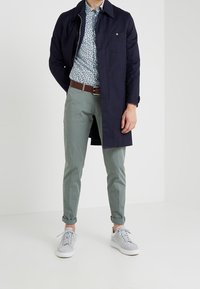 Cappotto blu navy, camicia a fantasia floreale, pantaloni verde chiaro e sneakers bianche. I pantaloni sono sartoriali con risvolti. L'outfit è casual e moderno.
