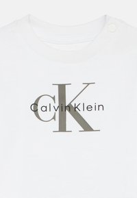 Valge puuvillane T-särk, millel on suur hall "CK" logo ja "CalvinKlein" tekst. Ümar kaelus, millel on nööbiga sulgemine õlal.
