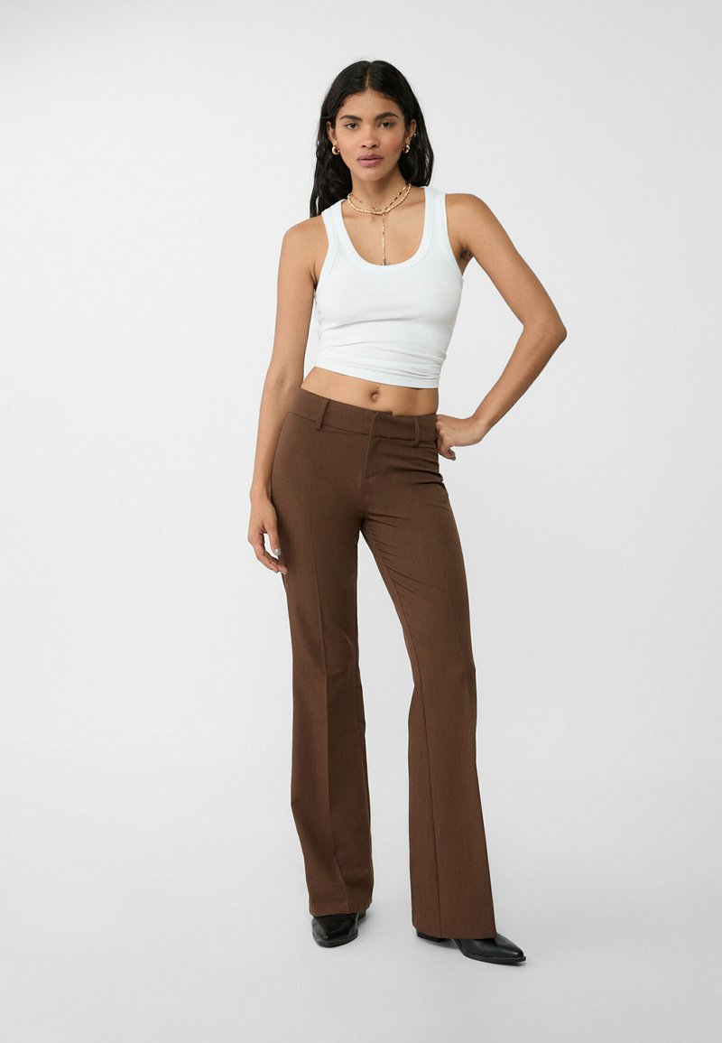 Pantaloni marroni a campana con una texture liscia e una vestibilità su misura, abbinati a un top bianco aderente. Il modello si trova su uno sfondo neutro.