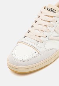 Hvit lær sneaker med beige semsket detaljer, perforert tå og beige snøring. Har gummisåle og subtil merking på tungen.