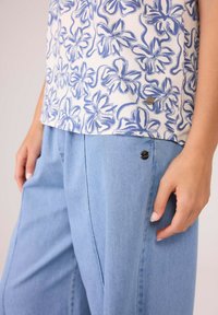 Leichtes, floral bedrucktes Oberteil aus creme- und blauen Stoff, kombiniert mit locker sitzenden hellblauen Jeanshosen, die über einen Knopfdetail am Bund verfügen.