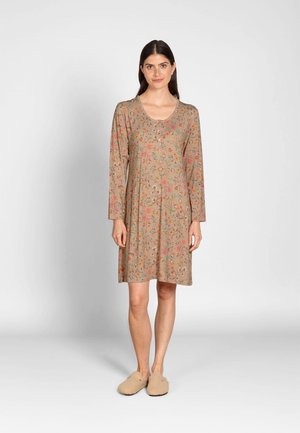 Vrouw staand, gekleed in een beige jurk met lange mouwen tot op de knie met bloemenpatronen en beige instappers op een effen achtergrond.