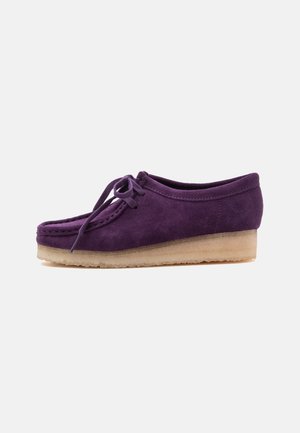 Zapato de ante morado con cordones, suela de crepé beige, costuras estilo mocasín y tacón bajo de cuña sobre fondo blanco.
