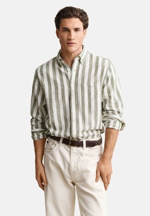 Mand iført grøn og hvid lodret striped skjorte med opkrogede ærmer, hvide bukser og et mørkt flettet bælte, stående mod en hvid baggrund.