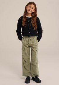 Jauna meitene ar gariem brūniem matiem, tērpusies melnā pogātā kardiganā, olīvzaļās kravas biksēs un melnās klumpē, stāv ar rokām kabatās.