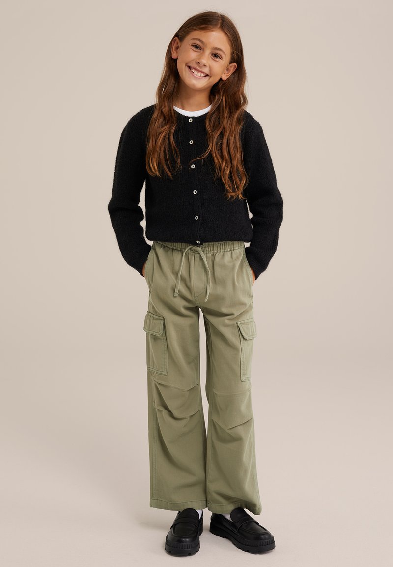 Jauna meitene ar gariem brūniem matiem, tērpusies melnā pogātā kardiganā, olīvzaļās kravas biksēs un melnās klumpē, stāv ar rokām kabatās.