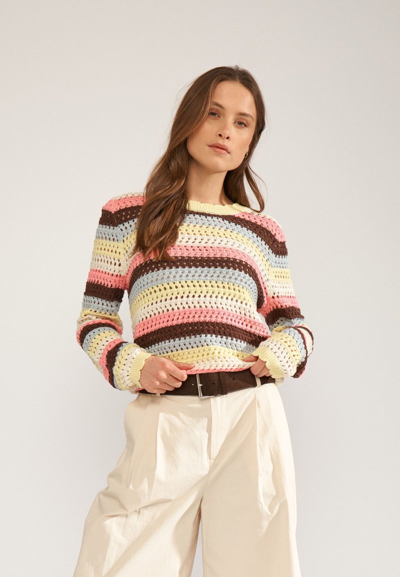 Jeune femme portant un pull en crochet rayé pastel et un pantalon large crème, posant les mains près de la ceinture sur un fond uni.