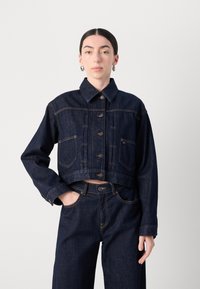 Kuttet denimjakke i mørk blå med krage, knappelukking og to frontlommer; har oransje søm og lange ermer.
