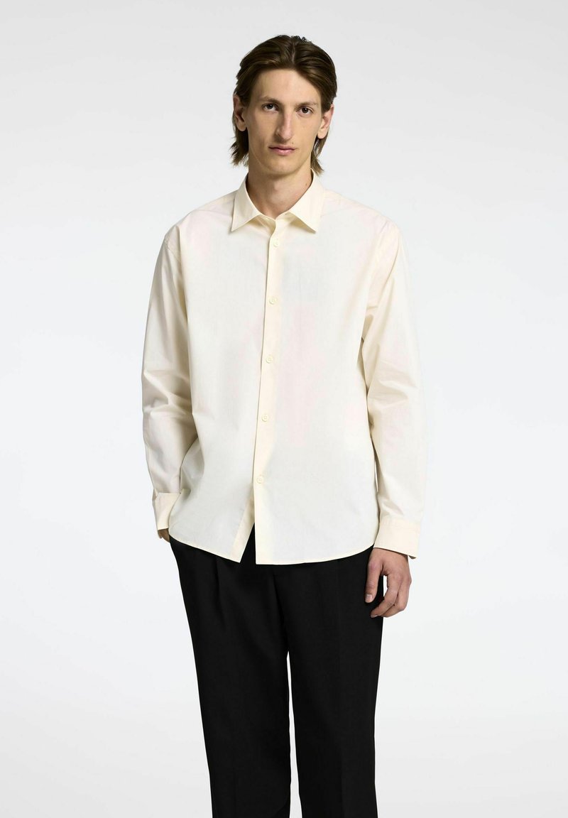 Camicia a maniche lunghe di un leggero color crema, con colletto button-down e placca frontale. Tessuto morbido con vestibilità rilassata, abbinata a pantaloni neri.