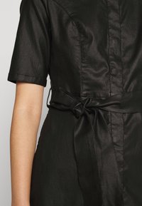 Czarna sukienka o błyszczącej satynowej powierzchni, z krótkimi rękawami, wiązaniem w talii oraz detalami szwów z przodu.